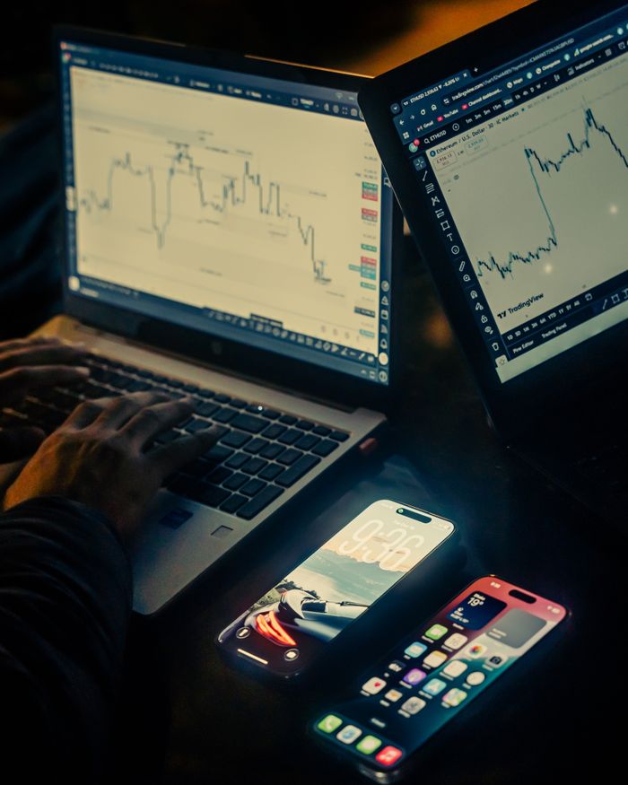 Offerings A trading workspace with laptops displaying charts and smartphones, set in a low light environment, Dhaka.
